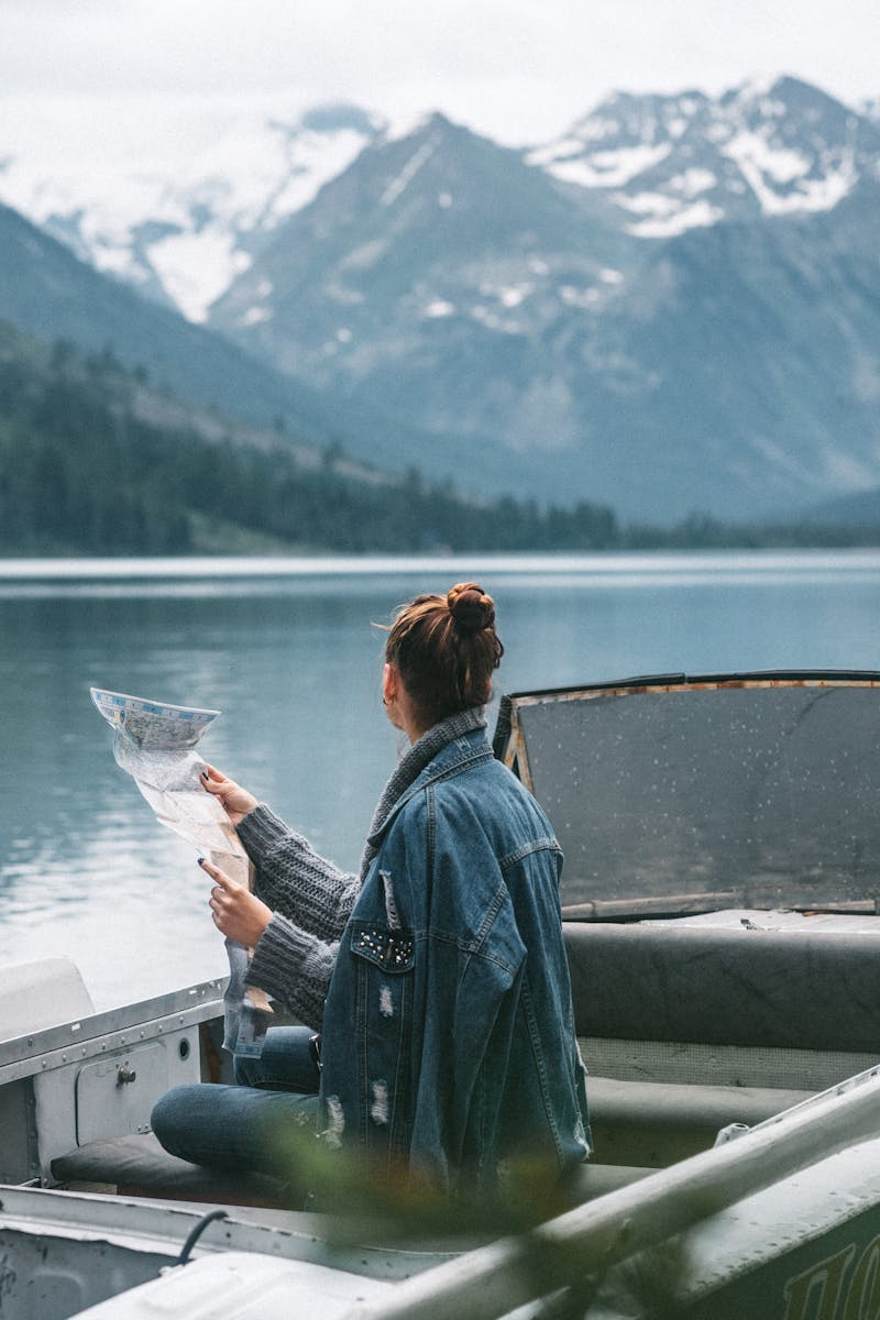 Side view of faceless female in warm clothes sitting with map in boat on lake while admiring view of snowy hills and woods in nature in daytime