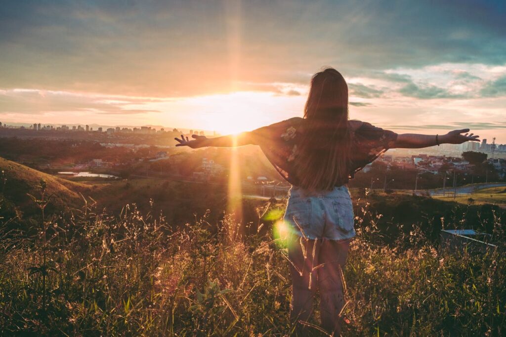 Woman with open arms enjoying sunset view over cityscape from a grassy hilltop.