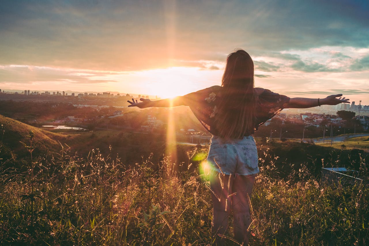 Woman with open arms enjoying sunset view over cityscape from a grassy hilltop.